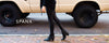 Person wearing black leggings and black ankle boots walking on a sidewalk with a beige vehicle in the background. The text