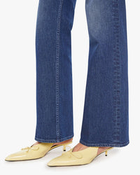 A person wearing Mother’s The Bookie Sneak in Perfectly Imperfect blue high-rise bootcut jeans and yellow pointed-toe low-heel shoes with small bows, standing on a white background.