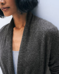 A woman with short, wavy black hair wears the White & Warren Cashmere Trapeze Cardigan in Dark Slate Heather over a light gray top, standing against a pale blue background. Her partially out-of-frame face highlights the cardigan's soft texture.
