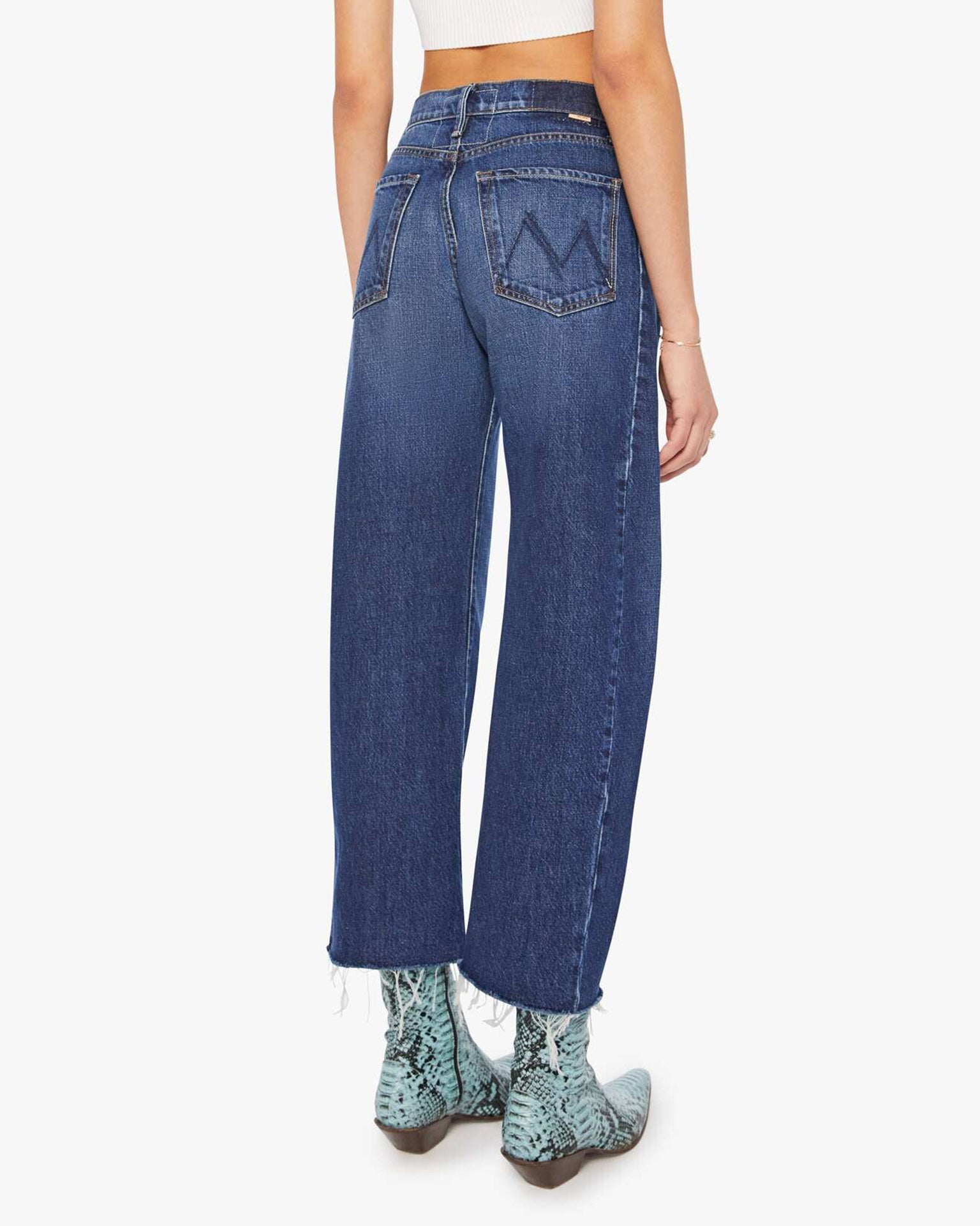A person is shown from the back wearing Mother’s Mid Rise Full-Pipe Flood Fray in Unplugged, blue barrel leg jeans with frayed hems, paired with a white crop top and light blue snakeskin boots. The jeans feature large back pockets with decorative stitching.