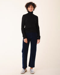 A person with short curly hair wears a black turtleneck, Prairie Underground’s Corduroy Carpenter Pant in Midnight, and silver shoes, standing against a plain light background.