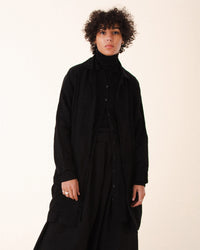 A person with curly hair wears the Prairie Underground Silk Habitual Coat in Black, a black turtleneck, and black pants, standing against a plain light background and looking calmly at the camera.