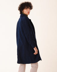 A person with short curly hair stands sideways, looking over their shoulder against a light background, wearing the Prairie Underground Silk Habitual Coat in Midnight over a green turtleneck and light pants.