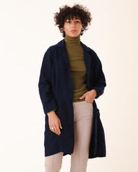 A person with short curly hair wears the Prairie Underground Silk Habitual Coat in Midnight over an olive green turtleneck and beige pants, standing against a plain light background.