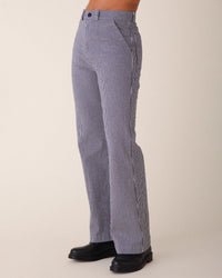 A person models Prairie Underground’s Long Smithy Pant in Oyster, with blue and white pinstripes, paired with black shoes against a plain beige background. The image focuses on the lower torso and legs.