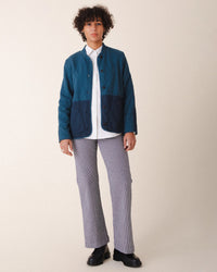 A person with short curly hair wears a Prairie Underground teal and navy quilted jacket over a white shirt, Long Smithy Pant in Oyster, and black shoes, standing against a plain beige background.
