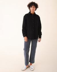 A person with short curly hair stands facing forward, wearing a black button-up shirt, Prairie Underground Corduroy Carpenter Pant in Cruiseliner, and silver slip-on shoes against a neutral background.