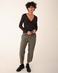 A person with short curly hair wears a fitted dark Prairie Underground V-neck long sleeve top, Prairie Underground Double Pleat Cords in Eucalyptus, and black shoes while standing against a plain light background.