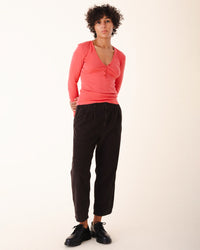 A person with curly short hair stands against a plain light background, wearing a coral long-sleeve top, Prairie Underground’s Double Pleat Cords in Brown Out, and black shoes, hands clasped behind their back.
