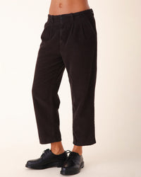 Someone stands against a plain light background wearing Prairie Underground's Double Pleat Cords in Brown Out—high-waisted, cropped, vintage-style dark brown corduroy trousers—paired with black lace-up shoes. Only the lower body is visible.