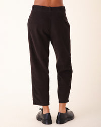 A person wears Prairie Underground's Double Pleat Cords in Brown Out with black oxford shoes, shown from behind against a plain light background. The pants' slightly cropped fit adds vintage flair by revealing the ankles.