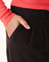 Wearing a coral long-sleeve, someone slips a hand into the pocket of Prairie Underground’s Double Pleat Cords in Brown Out, embodying a relaxed vintage style.