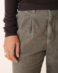 Wearing Prairie Underground's Double Pleat Cords in Eucalyptus and a long-sleeve dark brown top, the person stands with their left hand by their side, displaying a silver ring on their finger.