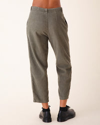 A person stands facing away, wearing Prairie Underground Double Pleat Cords in Eucalyptus—olive green, wide wale corduroy trousers with side pockets—paired with black socks and shoes. The background is plain and light-colored.