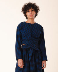 A person with short curly hair wears Prairie Underground's Twist Tie in Blue Mini Check, a cropped waist tie top, paired with a matching skirt made from deadstock flannel, standing against a plain light background.