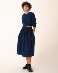 A person with short curly hair wears the Prairie Underground Twist Tie in Blue Mini Check, paired with black lace-up shoes, standing against a plain background.