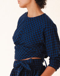 A person wears Prairie Underground’s Twist Tie in Blue Mini Check, a cropped top with a matching skirt in blue and black checkered deadstock flannel, shown from shoulders to mid-thigh against a plain light background, face partially out of frame.