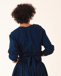 A person with short, curly hair is seen from behind wearing the Prairie Underground Twist Tie in Blue Mini Check, a long-sleeve flannel dress with a cropped waist tie and blue and black checks bow-tied at the waist.