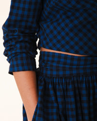 A person wears the Prairie Underground Twist Tie in Blue Mini Check— a long-sleeved, blue and black checkered top with a matching high-waisted skirt, one hand in the skirt pocket. The face is not visible.
