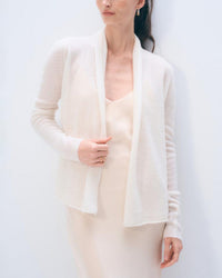 A woman in a White & Warren Cashmere Mini Trapeze cardigan in Soft White stands against a plain backdrop, holding the open sweater’s edge. Only her upper body and part of her face are shown.