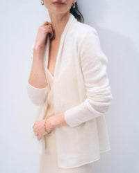 A woman wears the White & Warren Cashmere Mini Trapeze in Soft White over a light beige dress, paired with gold hoop earrings, a ring, and a bracelet. She stands against a plain light background, her face partially out of frame.