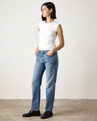 A woman stands against a plain wall, wearing the NSF Chloe Fitted Slub Jersey Muscle Tee in Soft White, blue jeans, and black shoes. Her hands are in her pockets as she looks to the side.
