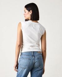A person stands facing away, wearing the NSF Chloe Fitted Slub Jersey Muscle Tee in Soft White and blue jeans against a plain, light-colored background.
