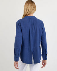 A person with long blonde hair is seen from behind, wearing the Bella Dahl Split Back Button Down in Midnight Surf and white pants, standing against a plain light background.
