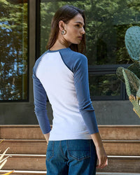 A person with long brown hair stands outdoors near a window and cactus, wearing the NSF Aria Fitted Baby Rib Raglan Tee in White/Indigo paired with blue jeans.