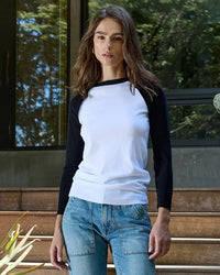 A woman with wavy brown hair wears the NSF Aria Fitted Baby Rib Raglan Tee in White/Black and blue jeans, standing outdoors by steps and a modern glass building, surrounded by greenery.