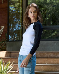 A person with long brown hair wears the NSF Aria Fitted Baby Rib Raglan Tee in White/Black and jeans while standing on wooden steps outside, looking over their shoulder with plants and a window in the background.