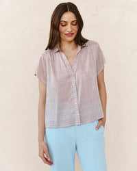 A woman with long brown hair wears the Bella Dahl Shirred Short Sleeve Shirt in Sunwashed Check and light blue pants, standing against a plain, light background.