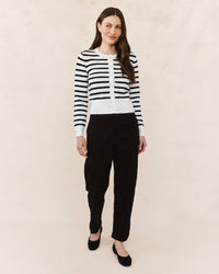 A woman with long brown hair and a slight smile poses against a neutral background, wearing the Bella Dahl Fitted Cardigan in Cheri Stripe, paired with black high-waisted pants and black flats.