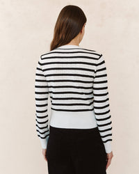 A woman with long brown hair stands facing away, wearing the Bella Dahl Fitted Cardigan in Cheri Stripe and black pants against a light beige background.