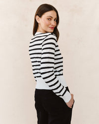 A woman with long brown hair, wearing the Bella Dahl Fitted Cardigan in Cheri Stripe and dark pants, stands slightly turned to the side, smiling over her shoulder against a light neutral background.
