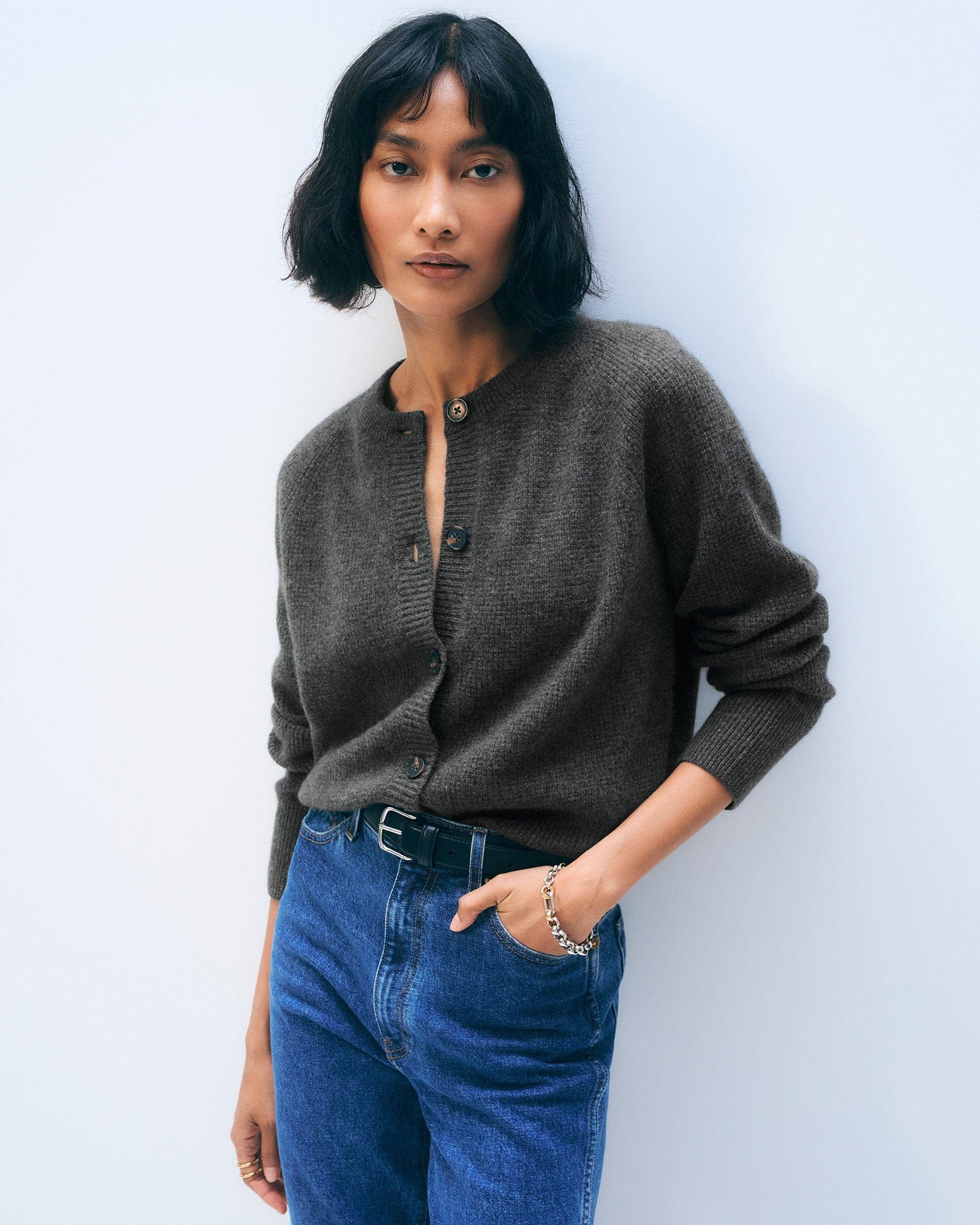 A woman with short dark hair wears the White & Warren Cashmere Waffle Cardigan in Dark Slate Heather, paired with blue jeans and a black belt. She stands against a plain light background, one hand in her pocket, looking confidently at the camera.