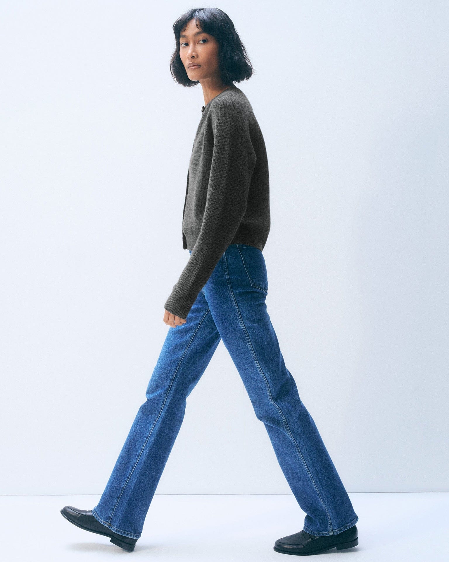 A woman with a short bob, in a White & Warren Cashmere Waffle Cardigan in Dark Slate Heather, blue straight-leg jeans, and black shoes, walks to the left against a plain light background.