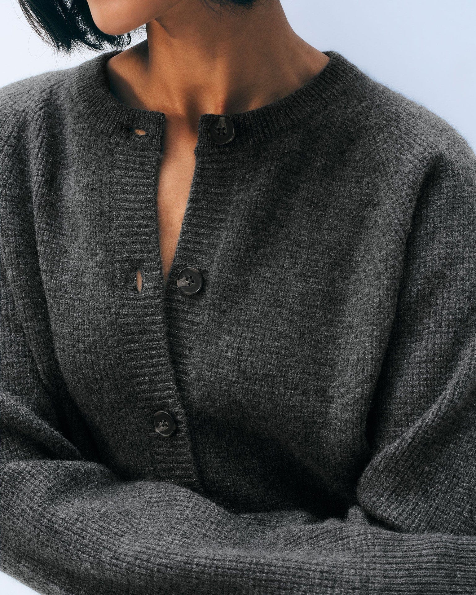 A person with short dark hair wears the White & Warren Cashmere Waffle Cardigan in Dark Slate Heather, buttoned up with one button undone, in front of a light blue background.
