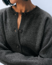 A person with short dark hair wears the White & Warren Cashmere Waffle Cardigan in Dark Slate Heather, buttoned up with one button undone, in front of a light blue background.