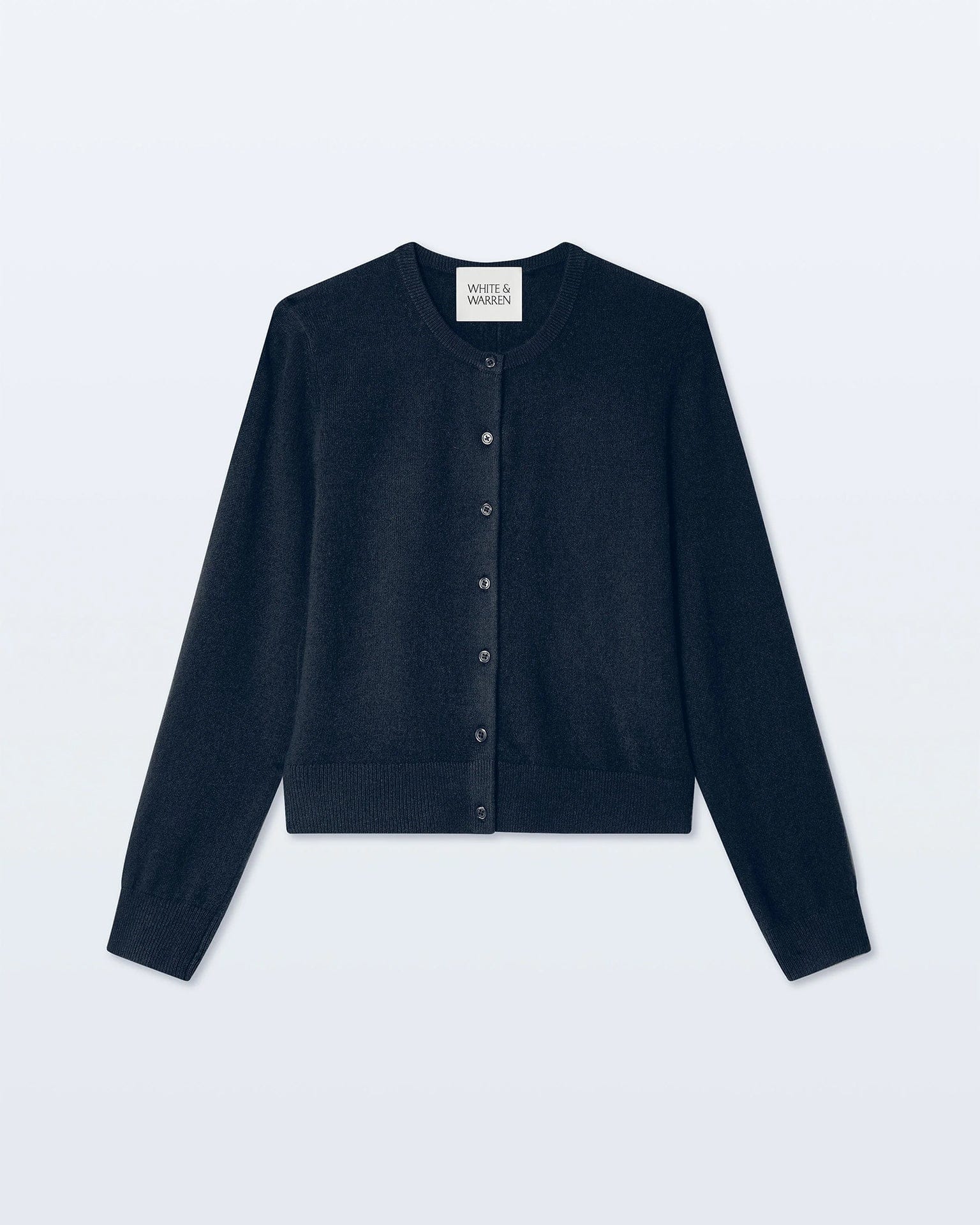 A White & Warren Cashmere Crewneck Cardigan in Deep Navy is laid flat on a light background, showing ribbed cuffs and hem, a button-up front, and an inner collar label.