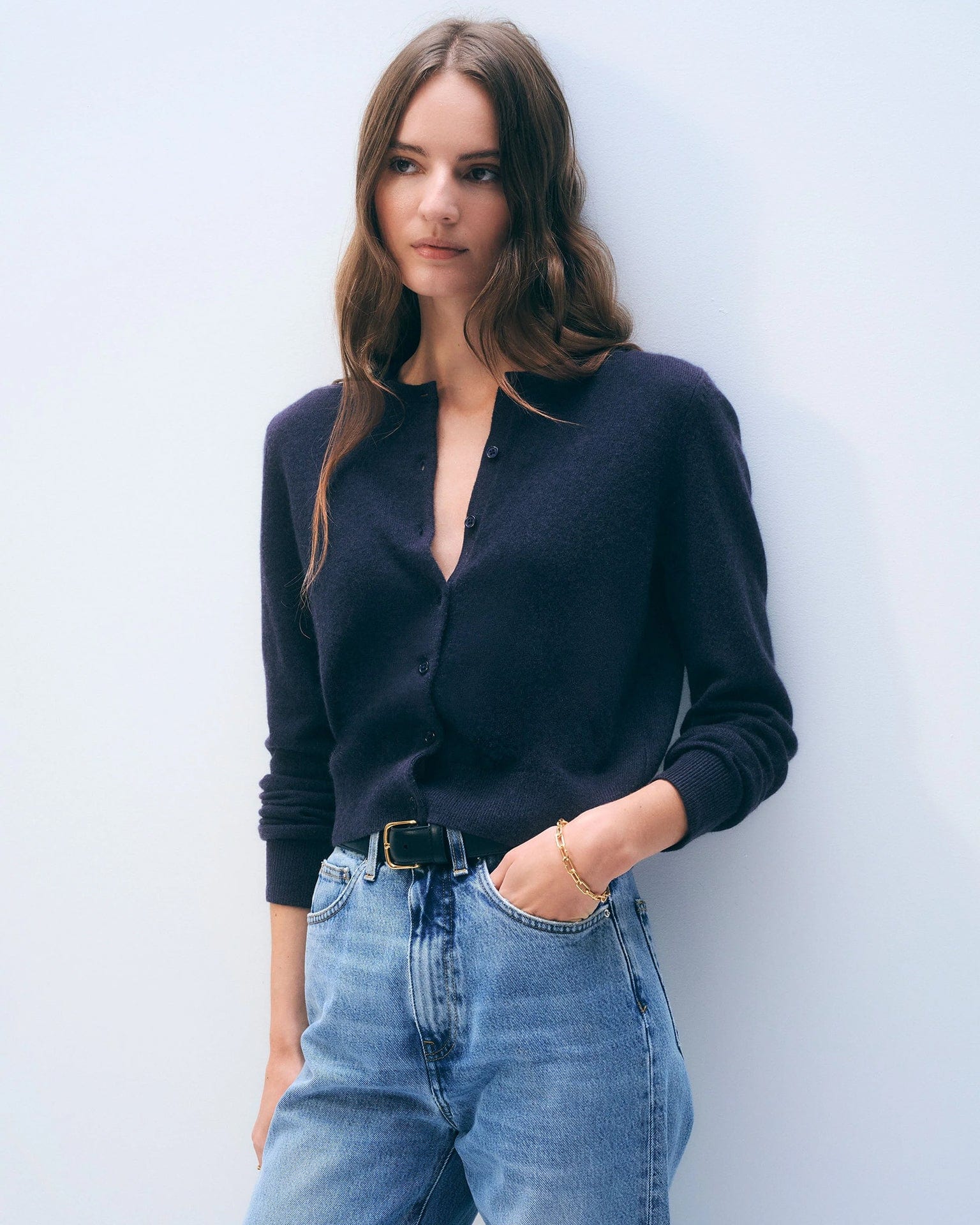 A woman with long brown hair wears a White & Warren Cashmere Crewneck Cardigan in Deep Navy, partially buttoned and tucked into high-waisted blue jeans with a black belt, standing against a plain light background.