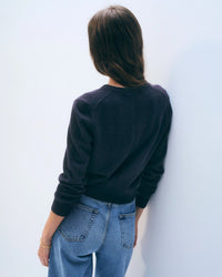 A person with long brown hair, wearing a White & Warren Cashmere Crewneck Cardigan in Deep Navy and blue jeans, stands facing a white wall, back to the camera.