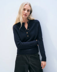 A woman with long blonde hair wears the White & Warren Cashmere Luxe Henley in Deep Navy and black pants, standing against a light blue background and looking at the camera with a relaxed expression.