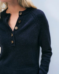 A person with long blonde hair wears the White & Warren Cashmere Luxe Henley in Deep Navy. The ribbed collar and five brown buttons are visible. The background is plain light blue, and the person’s face is partially out of frame.