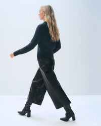 A woman with long blonde hair walks left against a plain light background, wearing White & Warren’s Cashmere Luxe Henley in Deep Navy paired with wide-leg black leather pants and black heeled boots.