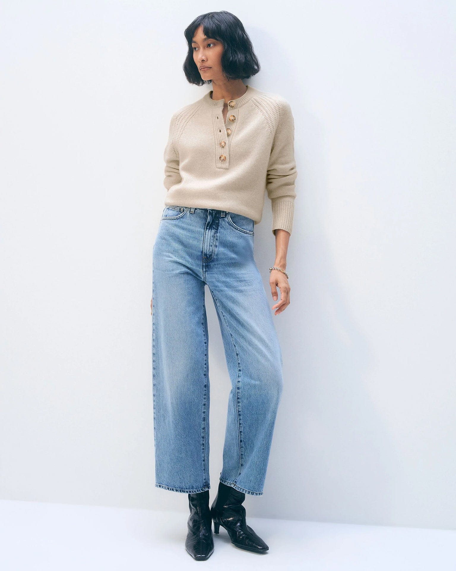 A woman with a short bob wears the White & Warren Cashmere Luxe Henley in Natural Heather, light blue wide-leg jeans, and black ankle boots, standing against a plain white background.