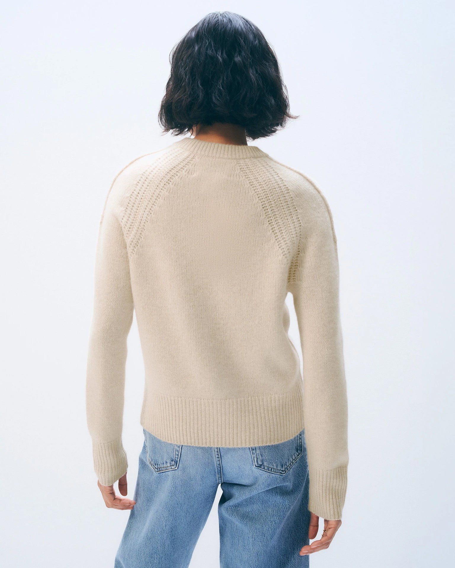 Person with short dark hair, seen from behind, wears the White & Warren Cashmere Luxe Henley in Natural Heather with light blue jeans, set against a plain, light background.