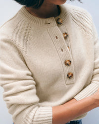 A person in the White & Warren Cashmere Luxe Henley in Natural Heather with a ribbed collar, stitched shoulders, and four large brown front buttons stands with arms crossed.