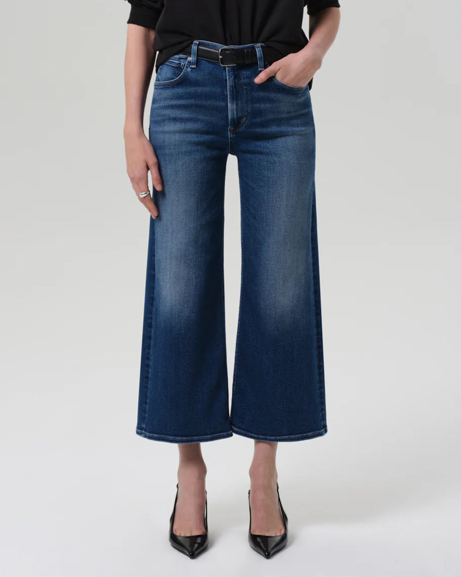 A person wears the Citizens of Humanity Lyra Wide Leg Crop in Augusta, styled with a black belt, top, and pointed-toe heels, standing with one hand in their pocket against a plain gray background.