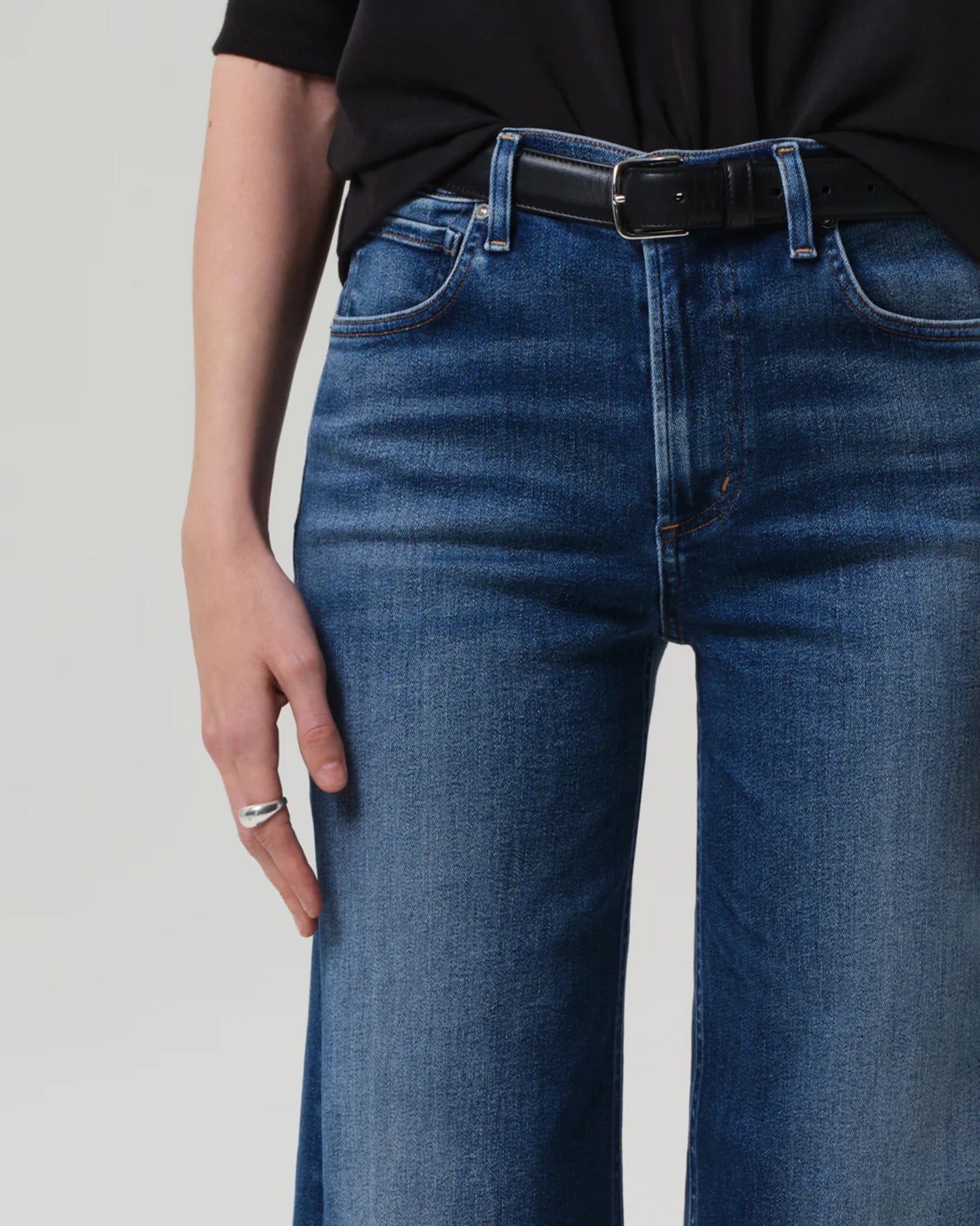A person wearing Citizens of Humanity's Lyra Wide Leg Crop in Augusta jeans, styled with a black belt and shirt. Their left hand with a silver ring rests by their side. The image is cropped at the waist and mid-thigh.